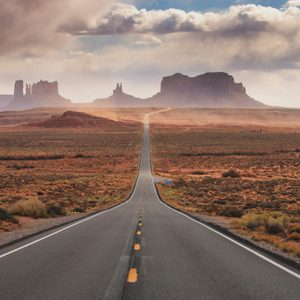 A straight road extends into vast desert landscape with distant rock formations under a cloudy sky.