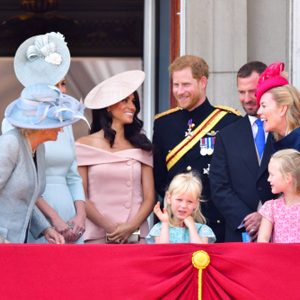 People stand smiling on a balcony, wearing formal attire and hats. Children are present, and a red railing with a gold emblem is visible.