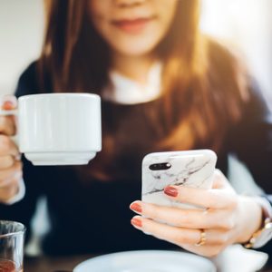 A person holds a white mug and a phone with a marble case, sitting at a table indoors, smiling softly.