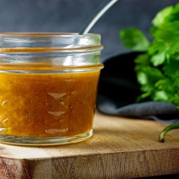 Jar of orange sauce sits on wooden board, surrounded by fresh cilantro and a green pepper, with a dark background.