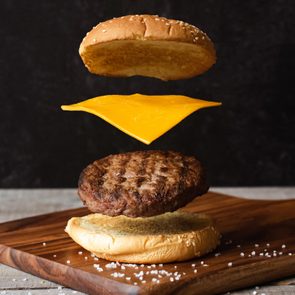 Burger ingredients float above a wooden board: sesame bun, cheese slice, patty, and bottom bun. Salt scattered around. Dark background.