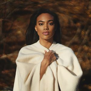 Person wrapped in a white shawl walks forward in a blurred forest background with autumn colors.