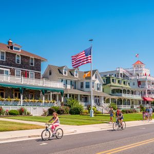 Bicyclists ride past colorful Victorian-style houses, with vibrant gardens and an American flag flying under a clear blue sky.