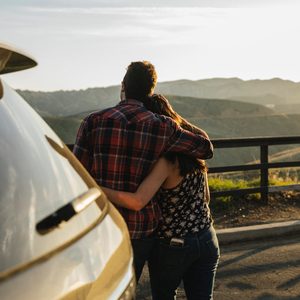 A couple embraces, gazing at mountainous scenery near a parked car at sunset.