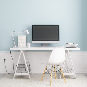 A computer sits idle on a minimalist desk with a lamp, books, and a chair in a light, airy room with a plant.