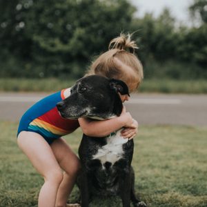 Child in colorful swimsuit hugs black dog on grassy area near a road, surrounded by a backdrop of dense greenery.