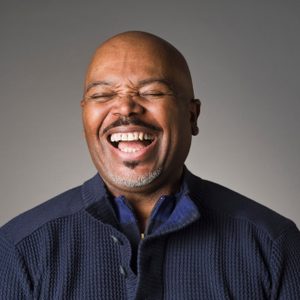 Man laughing with eyes closed, wearing a navy sweater, against a plain gray background.
