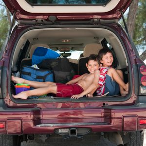 Two children relax in an open SUV trunk filled with beach gear, near a lakeside setting with trees and water visible in the background.