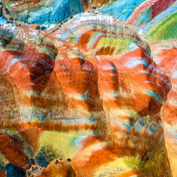 Colorful rock formations display vibrant layers; red, orange, blue, green, and yellow hues blend seamlessly, creating a surreal landscape from an aerial perspective.