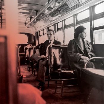 Woman sits calmly; man sits behind reading newspaper. They are on a nearly empty bus with large windows and advertisements on the ceiling.