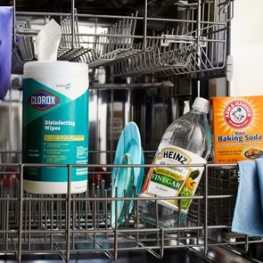 Cleaning supplies and a pair of gloves sit inside a dishwasher rack, including wipes, vinegar, baking soda, and a cloth in a kitchen.