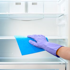 A gloved hand cleans a refrigerator shelf with a blue cloth in a white, light-filled kitchen environment.