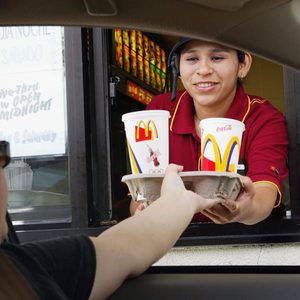 Drive-thru cashier hands beverage tray to car customer at fast-food window. Cups display McDonald's and Coca-Cola logos.
