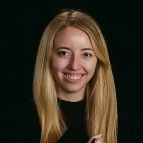 A woman smiles warmly, her long blonde hair framing her face against a plain black backdrop.