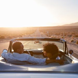Convertible drives into sunset on open road, with couples chatting and arm around each other, surrounded by desert landscape.