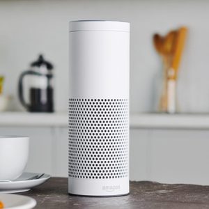 A cylindrical smart speaker stands on a kitchen counter, near a cup and saucer. A blurred background features utensils and a fruit bowl.