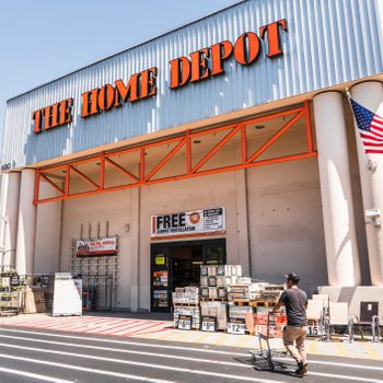 Storefront displays "The Home Depot," shopper pushes cart outside, flags and products line the exterior under a clear blue sky.