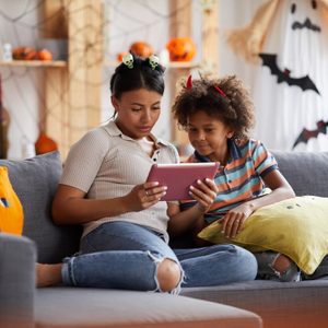 Two people, adorned in Halloween-themed headbands, sit together on a couch, engrossed in a tablet, surrounded by autumnal and festive decorations.