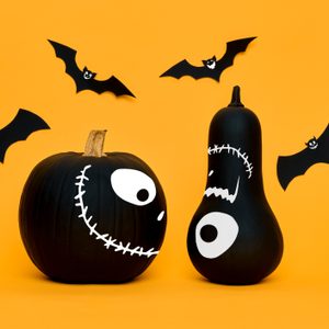 Black pumpkin and gourd painted with eyes and mouths, surrounded by five paper bats, against an orange background.