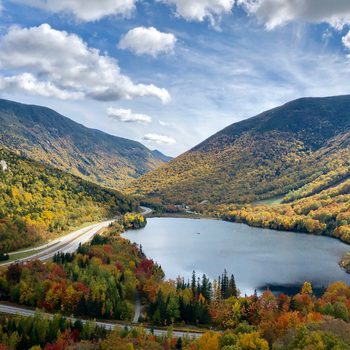 Lake surrounded by colorful autumn trees, nestled between mountains under a partly cloudy sky. A winding road splits the vibrant landscape.