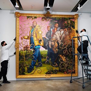 Two men in white coveralls adjust a large, ornate painting in a gallery setting, using a ladder for support.