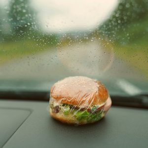 Burger wrapped in plastic sits on a car dashboard, with raindrops on the windshield and a blurred, rainy road outside.