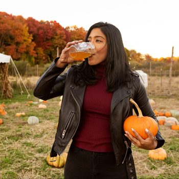 A person drinks from a glass jar, holding a pumpkin, surrounded by scattered pumpkins in a field with autumn foliage.
