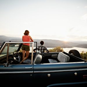 Two people stand in a convertible, gazing at a sunset over a scenic landscape with hills and a body of water.
