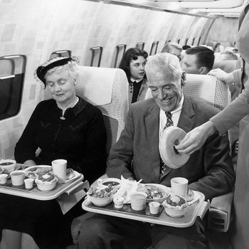 Passengers sit eating meals, served by a flight attendant on an airplane. The cabin is filled, and the atmosphere appears relaxed and pleasant.