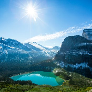 A bright sun illuminates snow-capped mountains surrounding a turquoise lake, with green forest at the base, under a clear blue sky.
