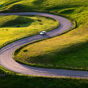 A white car navigates a winding road through lush, green hills under late afternoon sunlight.