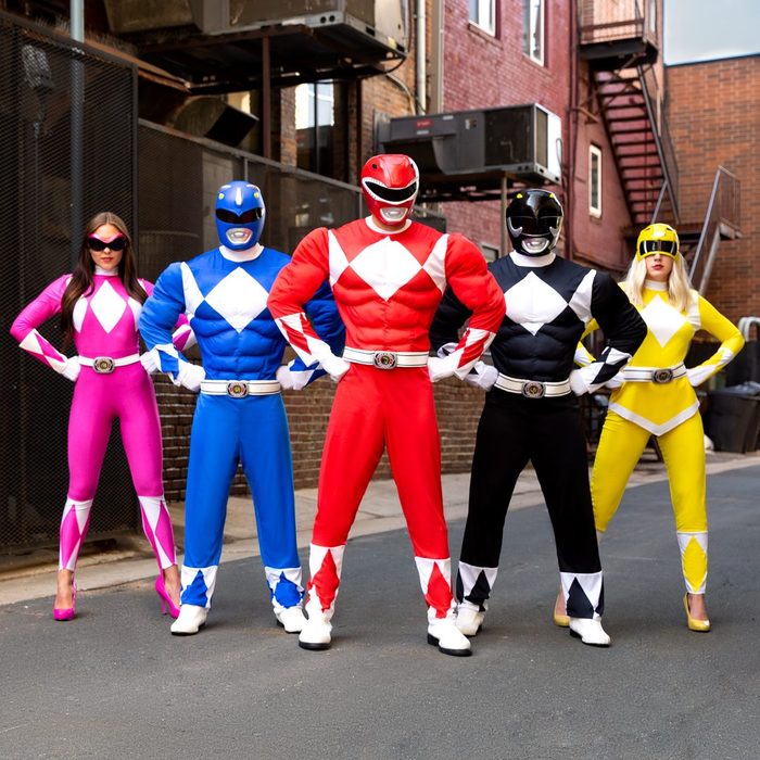 Five people in colorful superhero costumes stand confidently in an urban alleyway, surrounded by brick buildings and industrial structures.