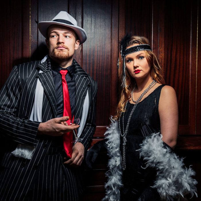 Man holds drink, wearing pinstripe suit and hat; woman stands beside, dressed in black flapper attire with feathered accessories, against wooden-paneled backdrop.
