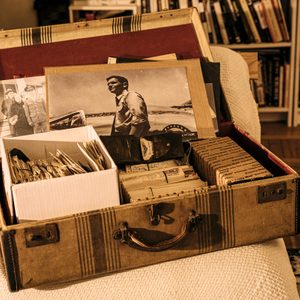 An open suitcase holds vintage photographs, letters, and keepsakes, resting on a bed in a cozy room with bookshelves.