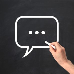 Hand holds chalk, drawing a speech bubble with ellipsis on a blackboard.