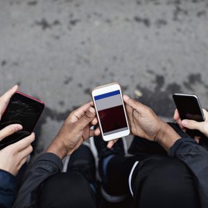 Three people holding smartphones, displaying screens, seated outdoors on pavement.