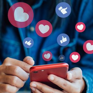 Hands hold a red phone, typing, with heart and thumbs-up icons floating around, against a blurred blue background.
