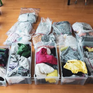 Plastic bins hold colorful folded clothes, arranged neatly on a wooden floor beside potted plants, with vacuum-sealed bags of clothes behind.