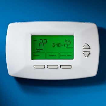 Thermostat displays 72 degrees, showing time 6:48 PM, set to heat, mounted on a blue wall with surrounding control buttons.