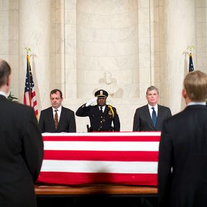 A casket draped with the American flag, flanked by solemn individuals, stands in a grand marble hall with statues and flags in the background.
