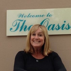 Smiling woman stands before a beige wall with a sign reading "Welcome to The Oasis."