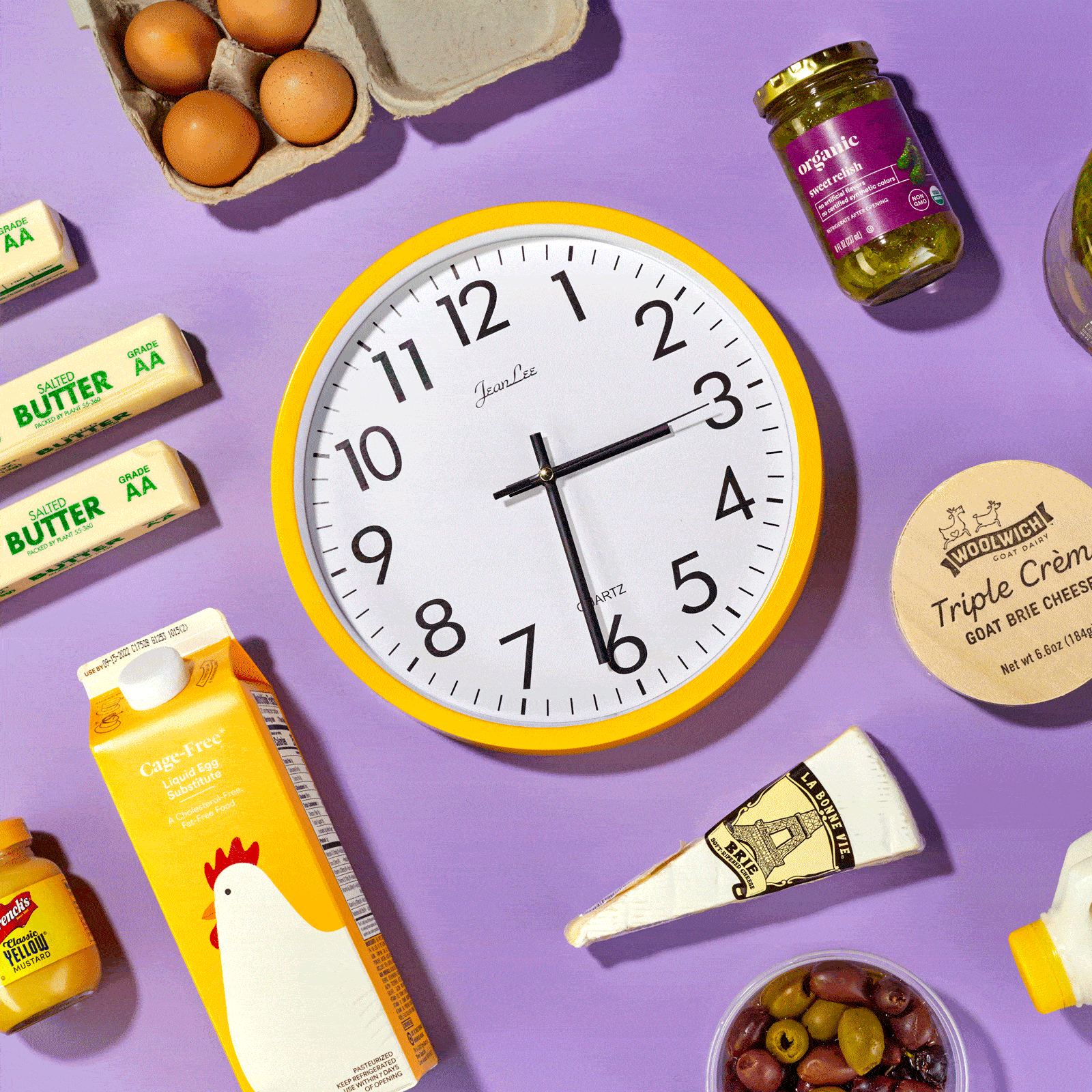 A clock shows different times surrounded by various food items including eggs, butter, relish, and cheese on a purple surface.