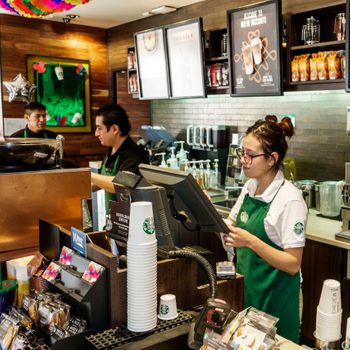 Barista working at a coffee shop counter with stacked cups, payment device, and menu boards; colleagues prepare drinks in the background.