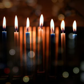 Lit candles flicker gently against a dark background, surrounded by soft, out-of-focus light orbs.