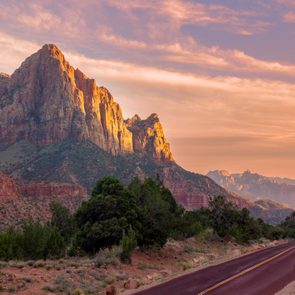 Sunlit mountains rise dramatically, basking under a vibrant sky, adjacent to a winding road and lush greenery.