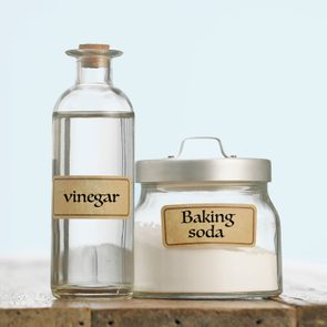 A vinegar bottle stands beside a baking soda jar on a wooden surface against a plain light background.