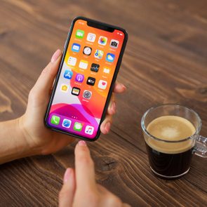 A smartphone displays colorful app icons; a finger gestures on it. Nearby, a glass cup holds dark coffee on a wooden table.