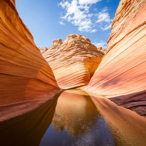 Sandstone canyon reflects in still water under a bright blue sky with wispy clouds, showcasing curved, multicolored rock formations.