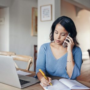 A person writes in a notebook while on the phone, sitting at a table with a laptop in a home setting.