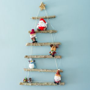 A wall-hung Christmas tree made of sticks features Santa, snowman plushies, and decorations. It's displayed on a plain blue wall.
